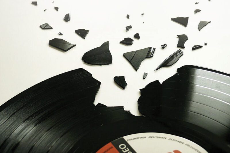 A black vinyl record lies broken, with several jagged pieces scattered around it on a white surface.
