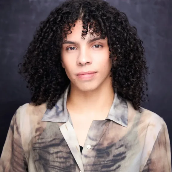 Person with medium-length curly hair and a nose ring, wearing a semi-sheer, patterned button-up shirt, posed in front of a dark background.