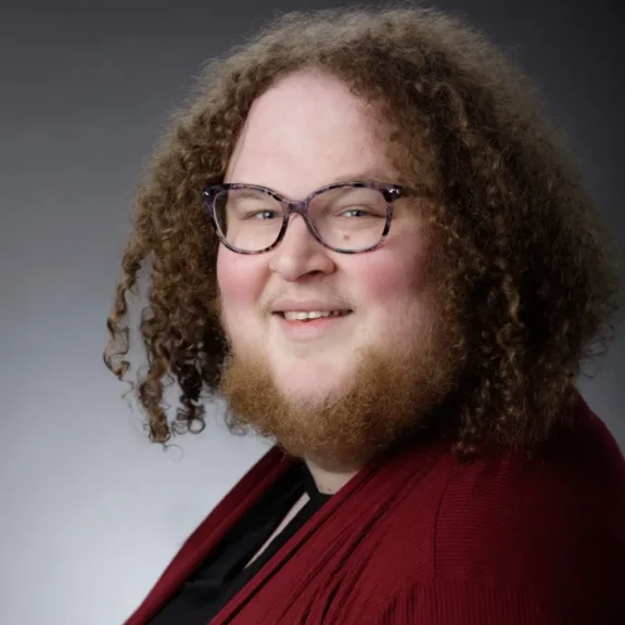 A person with curly hair, glasses, and a beard is smiling while wearing a red sweater and black shirt against a gray background.