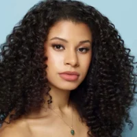 A woman with long, voluminous, curly dark hair and natural makeup poses against a light blue background, wearing a necklace with a green pendant.