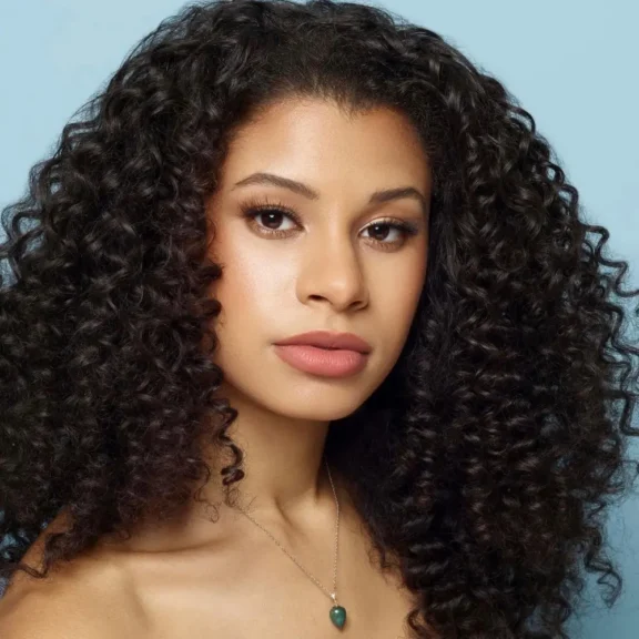A woman with long, voluminous, curly dark hair and natural makeup poses against a light blue background, wearing a necklace with a green pendant.