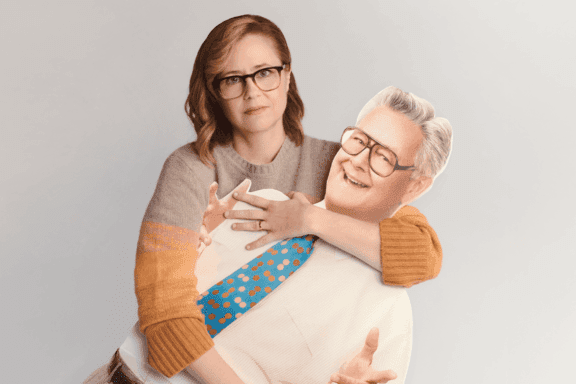 A woman with glasses holds a life-size cardboard cutout of an older man wearing glasses, a white shirt, and a colorful polka dot tie.
