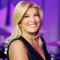 A smiling woman with straight blond hair wearing a sleeveless black top stands in front of a blurred purple background.