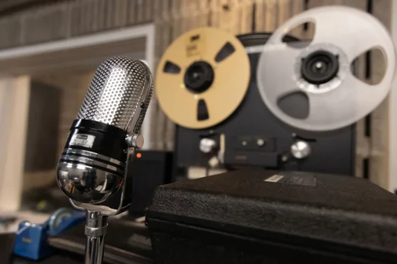 A vintage silver microphone is positioned in front of a reel-to-reel tape recorder in a recording studio.