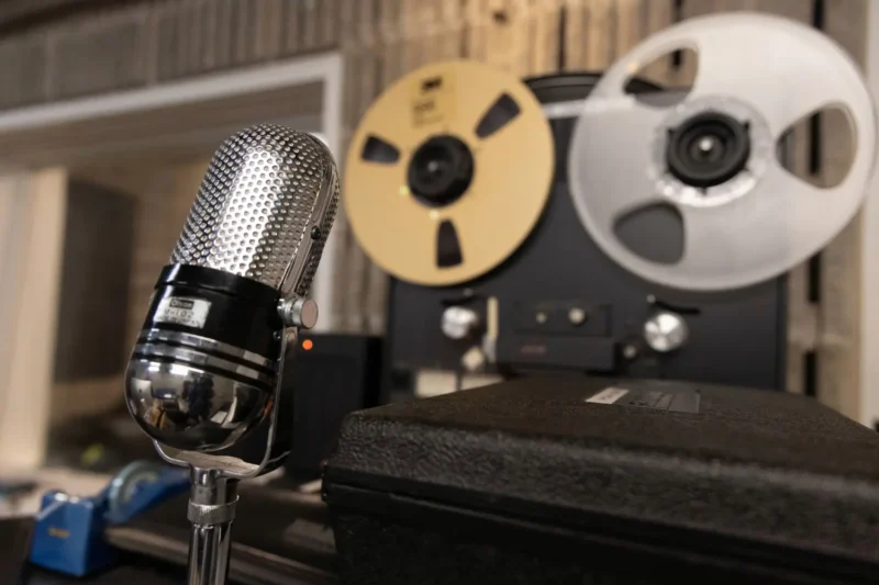 A vintage silver microphone is positioned in front of a reel-to-reel tape recorder in a recording studio.
