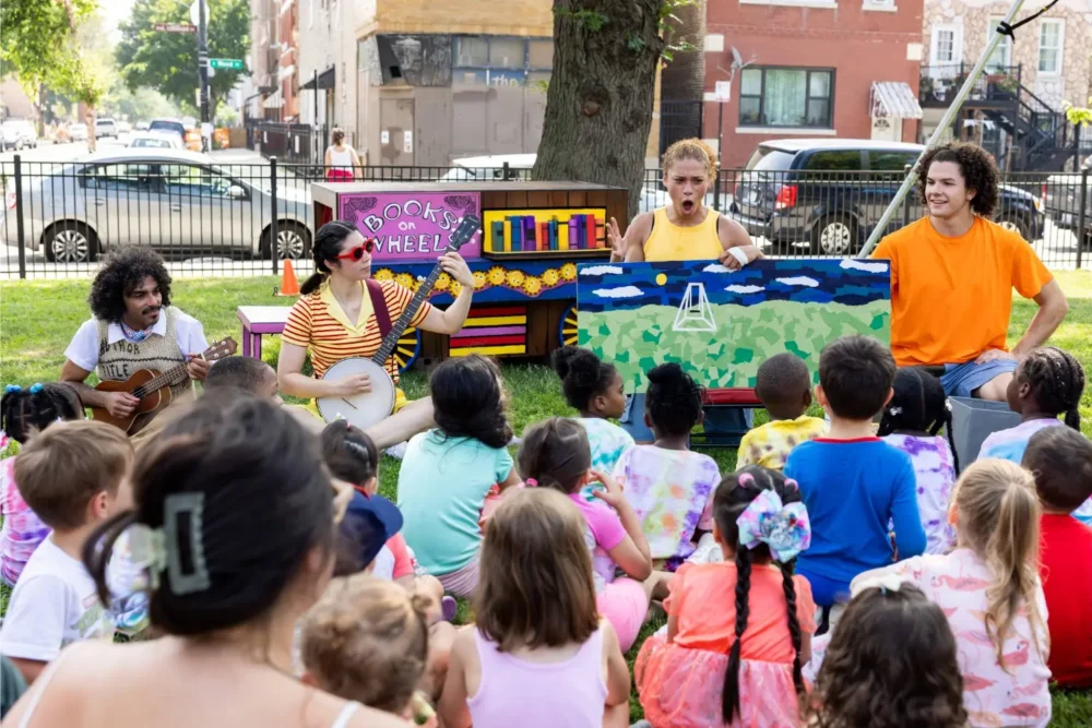 A group of performers entertain a crowd of children outdoors with music and storytelling, using colorful props and instruments on a grassy area.