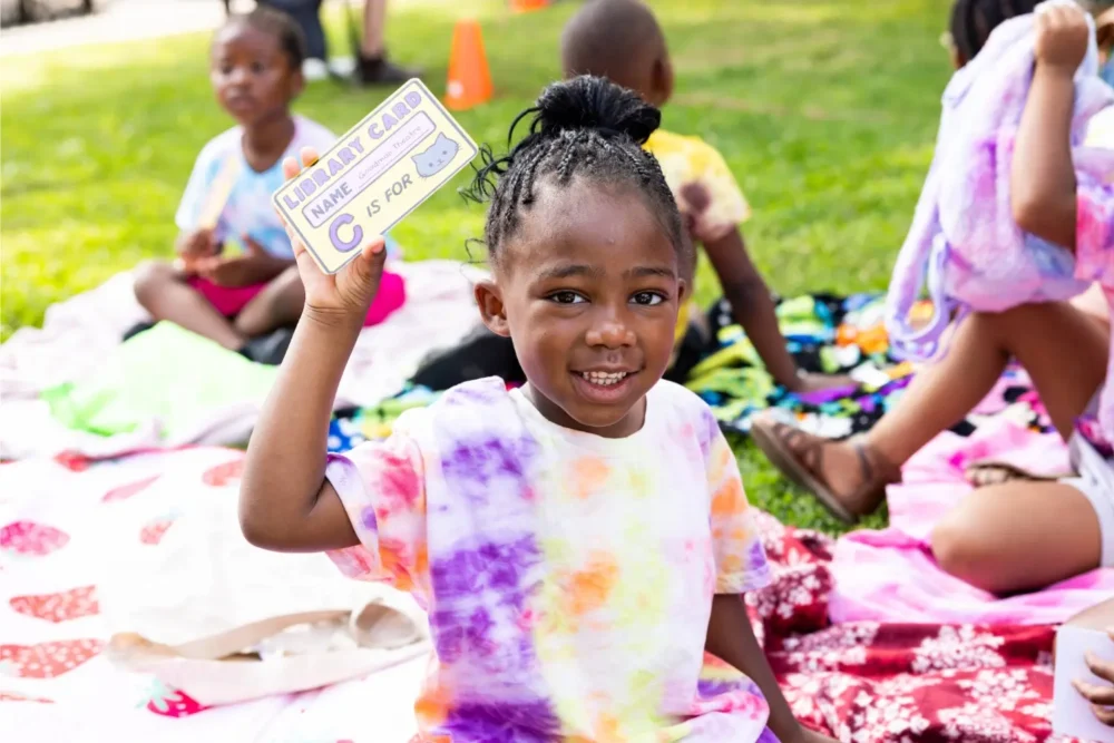 A young child sitting on a blanket outdoors holds up a card that says "Library Card" and "C is for" while other children sit in the background.