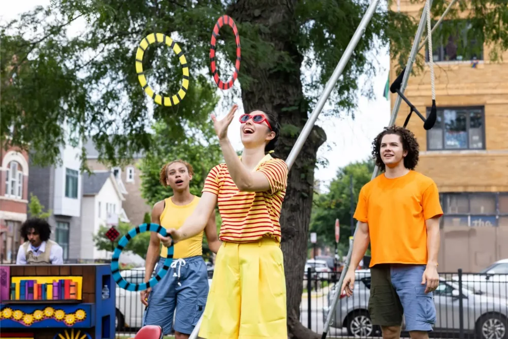 Three people outdoors watch as a person in a striped shirt and yellow shorts juggles colorful rings. Trees and buildings are visible in the background.