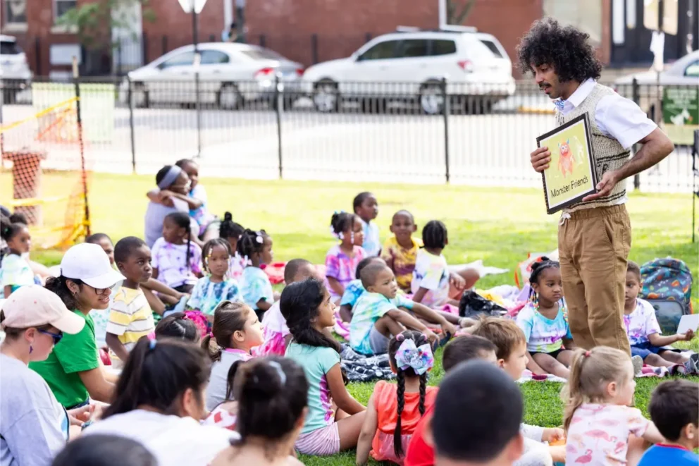An adult stands and reads from a book to a group of young children sitting on the grass in a park, with cars and buildings visible in the background.