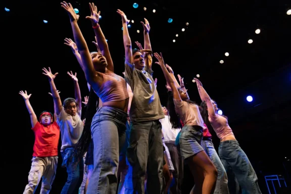 A group of people on stage stand close together, raising their arms upwards under bright stage lights, performing a dance or theatrical routine.