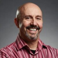 Bald man with a goatee and mustache wearing a red and white striped shirt, smiling with arms crossed against a gray background.