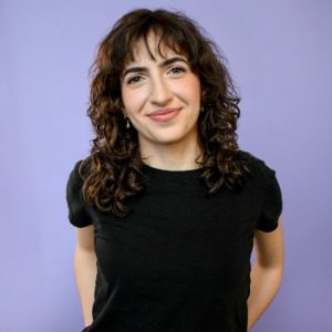 A person with curly brown hair wearing a black shirt stands in front of a plain light purple background, smiling slightly at the camera.