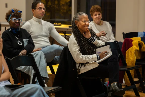 A group of adults sit on black chairs indoors, listening attentively; one woman in front is smiling and holding a notebook and pen.