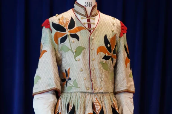 A mannequin displays an ornate, embroidered costume with colorful floral patterns, gold buttons, and red accents on the shoulders, set against a dark blue backdrop.