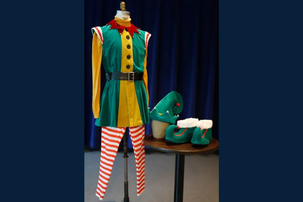 A mannequin displays a colorful elf costume with a green and yellow tunic, red and white striped leggings, and a black belt. Matching green hat and shoes sit on a nearby table.