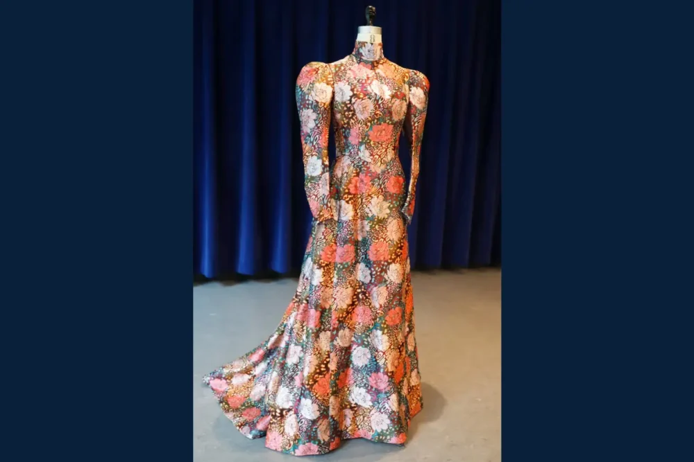 A long-sleeved, floor-length gown with a high neckline featuring a colorful floral pattern, displayed on a dress form against a dark blue curtain backdrop.