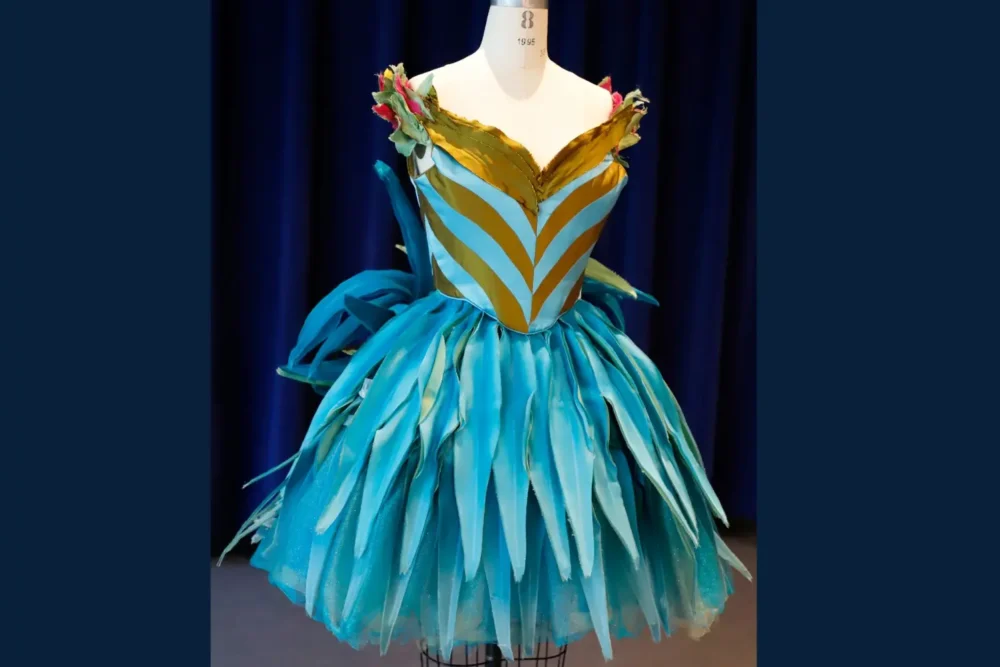A mannequin displays a vibrant blue and gold dress with a leaf-like layered skirt and floral accents on the neckline, set against a dark blue curtain background.