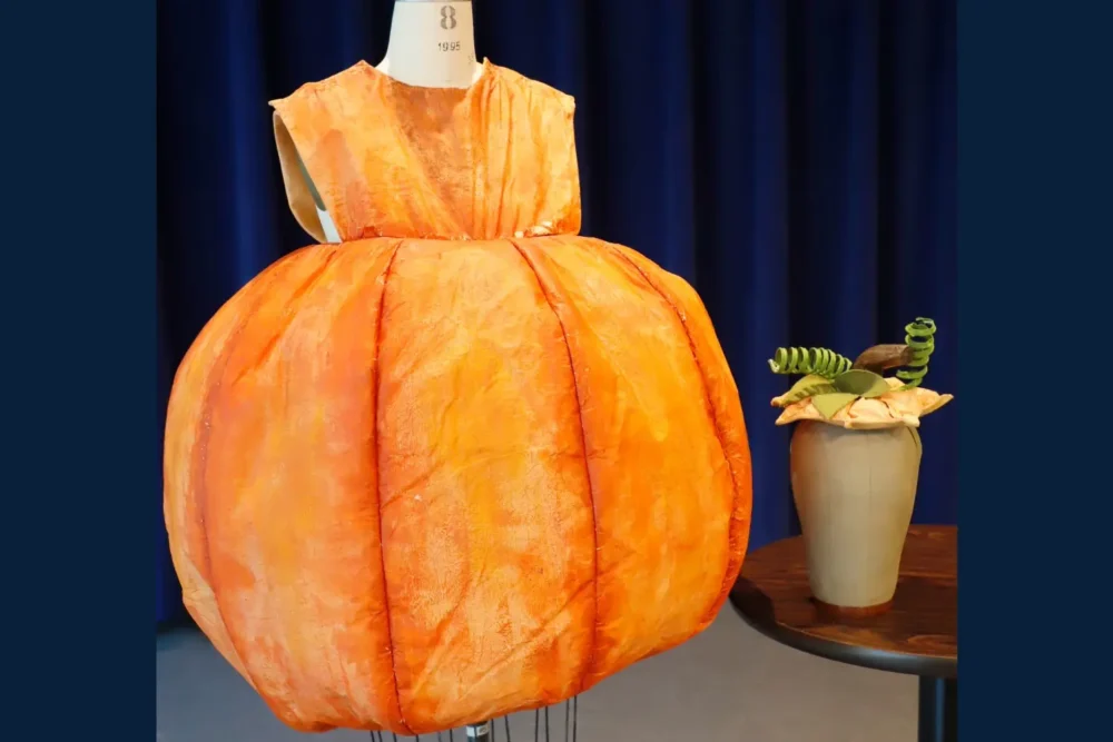 A mannequin displays an orange pumpkin-shaped dress next to a vase with a pumpkin-themed arrangement on a wooden table, against a dark blue curtain backdrop.