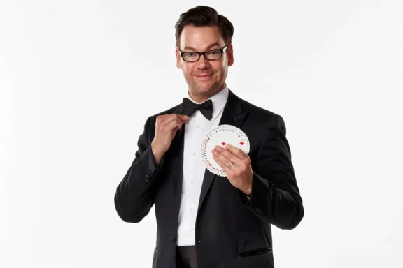 A man in a tuxedo adjusts his bow tie with one hand and holds a fanned-out deck of playing cards in the other, standing against a plain white background.