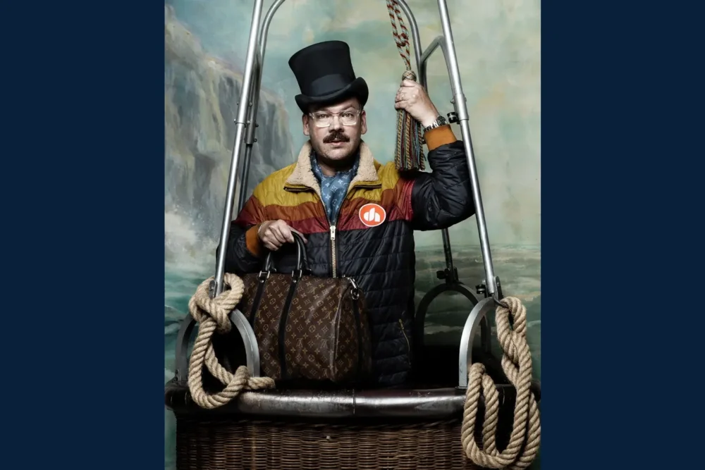 A man wearing a top hat and colorful jacket stands in a hot air balloon basket, holding a rope and a brown patterned bag, with a painted backdrop behind him.