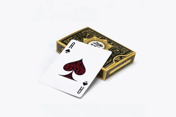 A single ace of spades playing card rests on top of a closed, ornate deck of cards with a black and gold design.