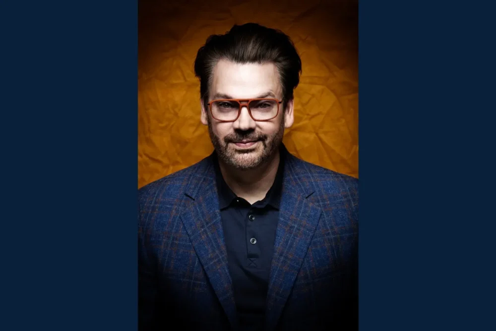 Man with dark hair, beard, and glasses wearing a blue plaid blazer and dark shirt, posed against a textured orange background.