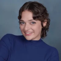 A person with short, curly brown hair and blue eyes smiles at the camera, wearing a blue turtleneck top against a plain blue background.