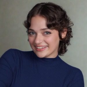 A young woman with short, curly brown hair and blue eyes smiles at the camera while wearing a dark blue, long-sleeve top against a neutral background.