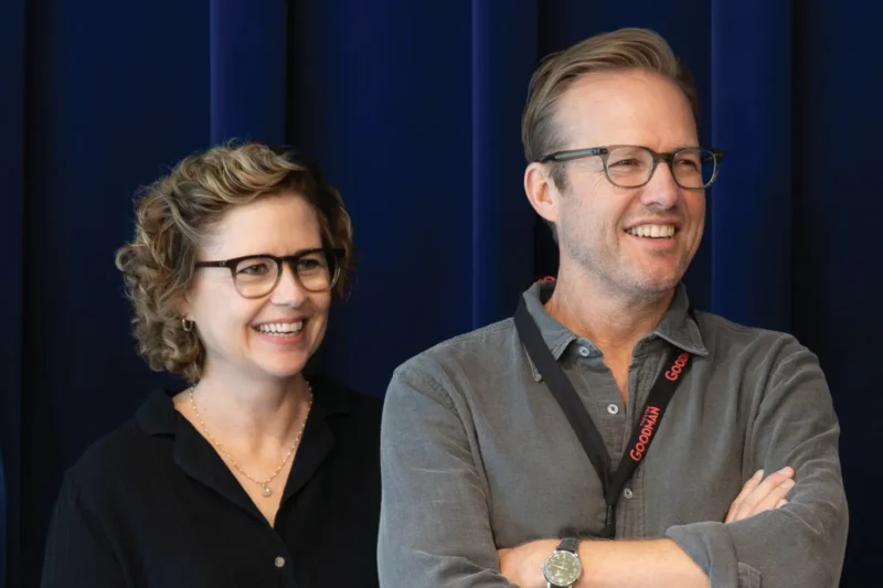 Two people wearing glasses and casual clothing stand side by side, smiling in front of a dark blue curtain.