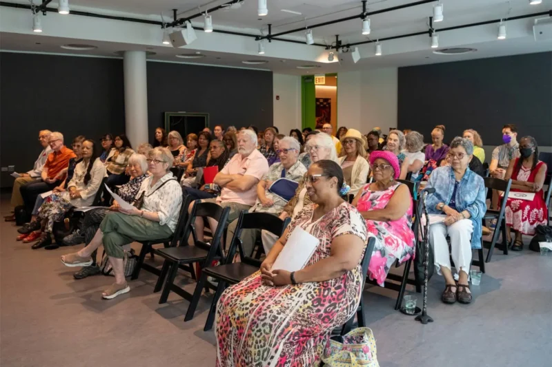 A diverse group of adults sit in rows of chairs, attentively watching a presentation in a well-lit room.