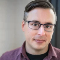 A person with short brown hair, glasses, and a maroon collared shirt looks toward the camera with a neutral expression. The background is softly blurred.
