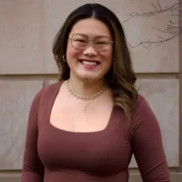 A person with long brown hair, glasses, and a necklace stands smiling in front of a beige stone wall, wearing a brown square-neck top.