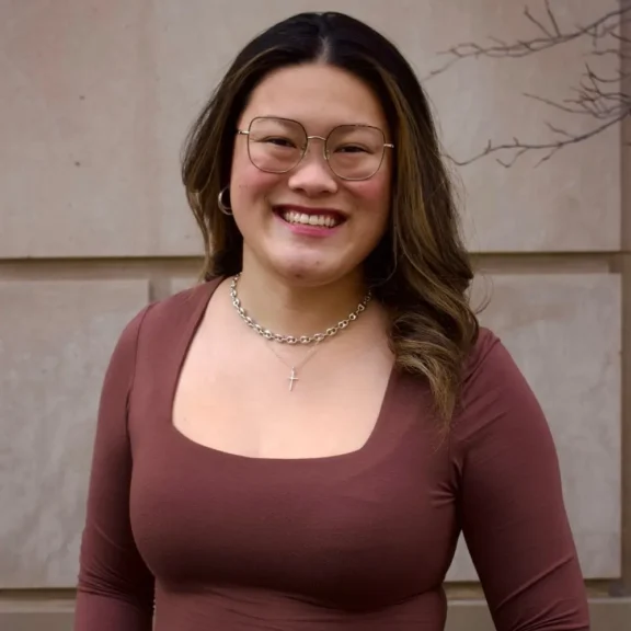 A person with long brown hair, glasses, and a necklace stands smiling in front of a beige stone wall, wearing a brown square-neck top.