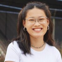 A young person with long dark hair, glasses, and a white shirt smiles outdoors.