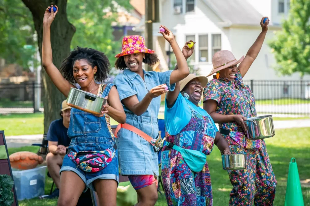 Four women outdoors, smiling and dancing while holding pots and shakers, with houses and trees visible in the background.