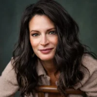 A woman with long, wavy dark hair and light eyes is seated, facing the camera, wearing a neutral-colored, textured jacket against a dark background.