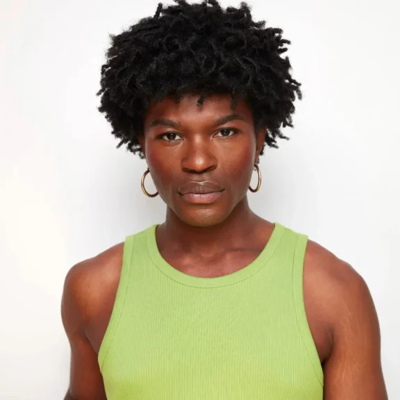 A person with short curly hair, wearing gold hoop earrings and a sleeveless light green top, stands against a plain white background.