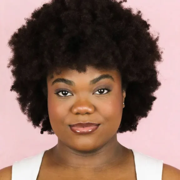 A person with natural, voluminous afro hair and a neutral facial expression stands against a pink background, wearing a white top.