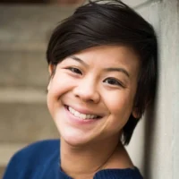 A person with short dark hair and a blue sweater smiles while leaning against a concrete wall, with blurred steps in the background.