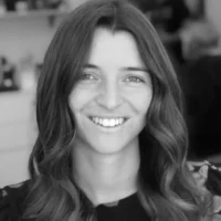 A woman with long, wavy hair smiles at the camera in a black and white photo, with a blurred indoor background.