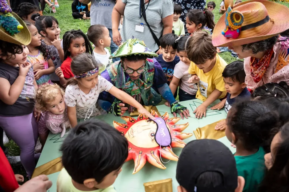 A group of children and adults gather around a sun-shaped board game outdoors, watching as a person in costume points to the game piece.