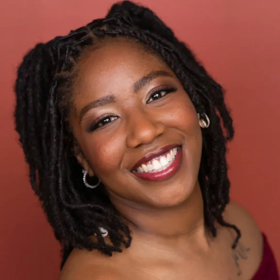 A woman with medium-length locs, wearing hoop earrings and off-shoulder clothing, smiles in front of a reddish-brown background.