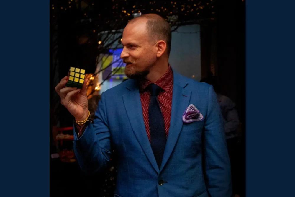 A man in a blue suit and red shirt holds a solved Rubik's Cube in his right hand, standing indoors with dim lighting and string lights in the background.