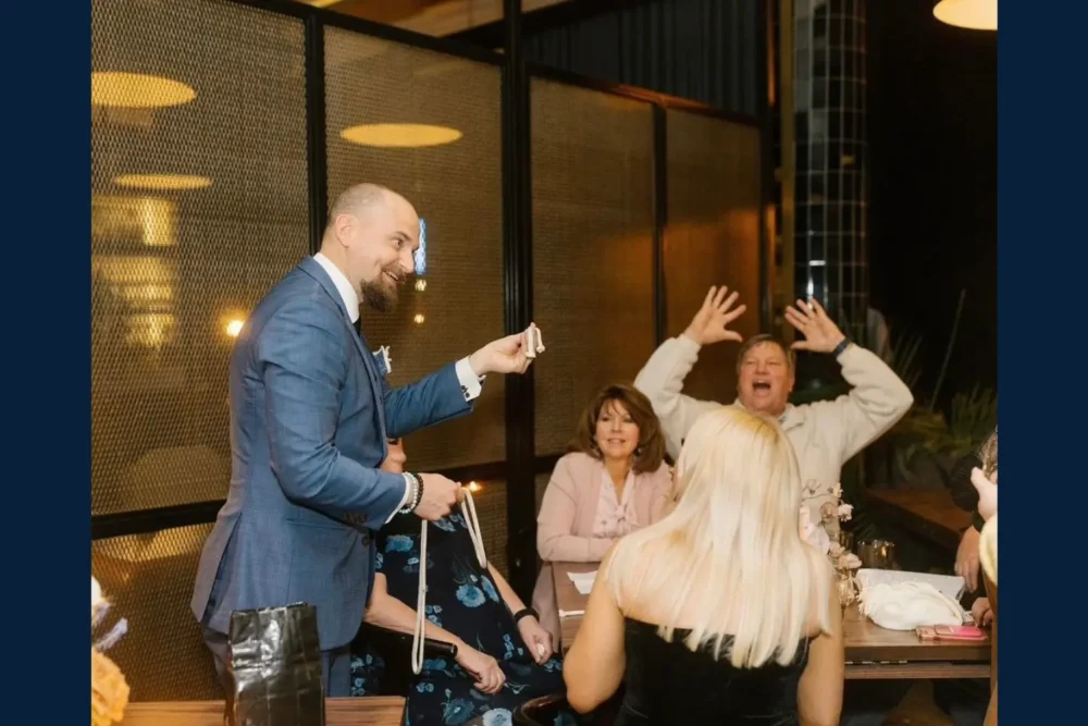 A man in a suit performs a magic trick with a rope for a group of people seated at a table; one man reacts with raised arms and an excited expression.