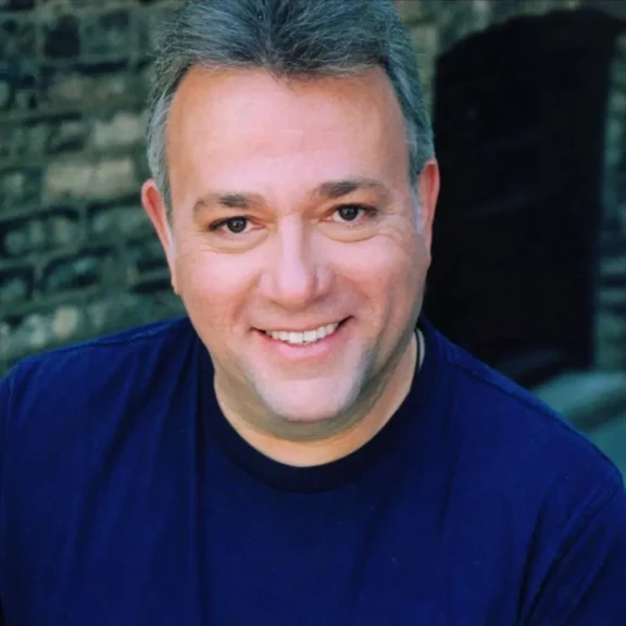 A man with short gray hair and a dark blue shirt smiles at the camera in front of a blurred brick background.