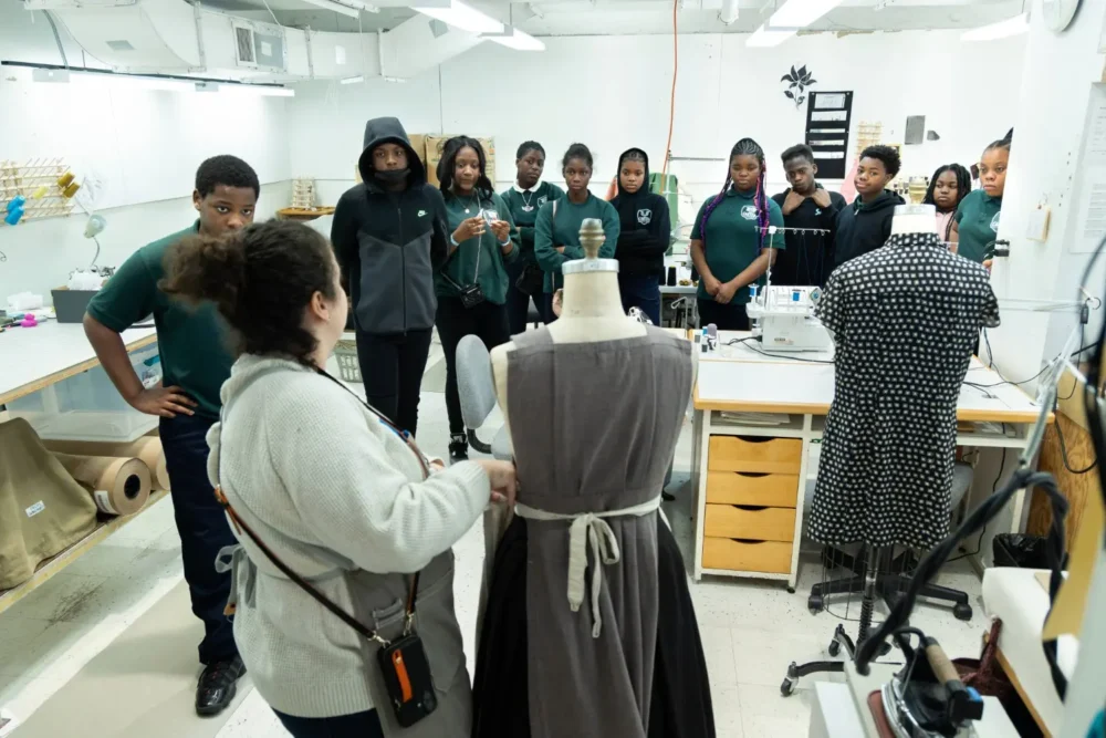 A group of students observes an instructor demonstrating dressmaking on a mannequin in a sewing classroom.