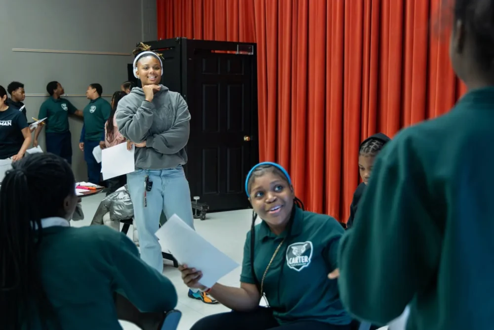 A group of students interact and hold papers in a classroom with red curtains; one person stands while others are seated and talking.