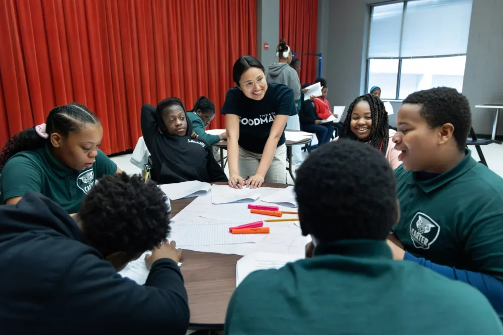 A group of students sits around a table working on assignments, while an adult stands and interacts with them in a classroom setting.