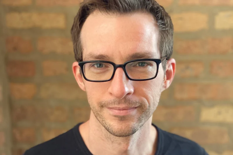 A man with short brown hair and black glasses is standing in front of a brick wall, wearing a black shirt and looking at the camera.