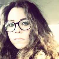 A woman with long, wavy brown hair and large glasses looks at the camera inside a car.